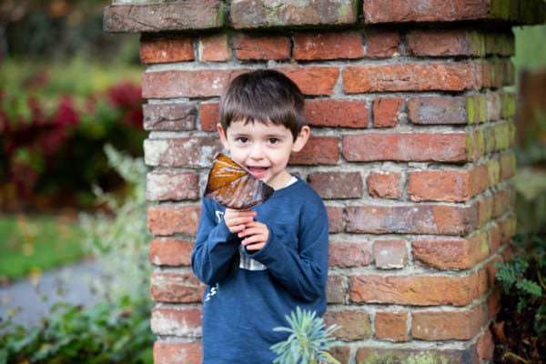 little boy holding a leaf and smiling against a brick wall at his autumn family photoshoot in Sevenoaks
