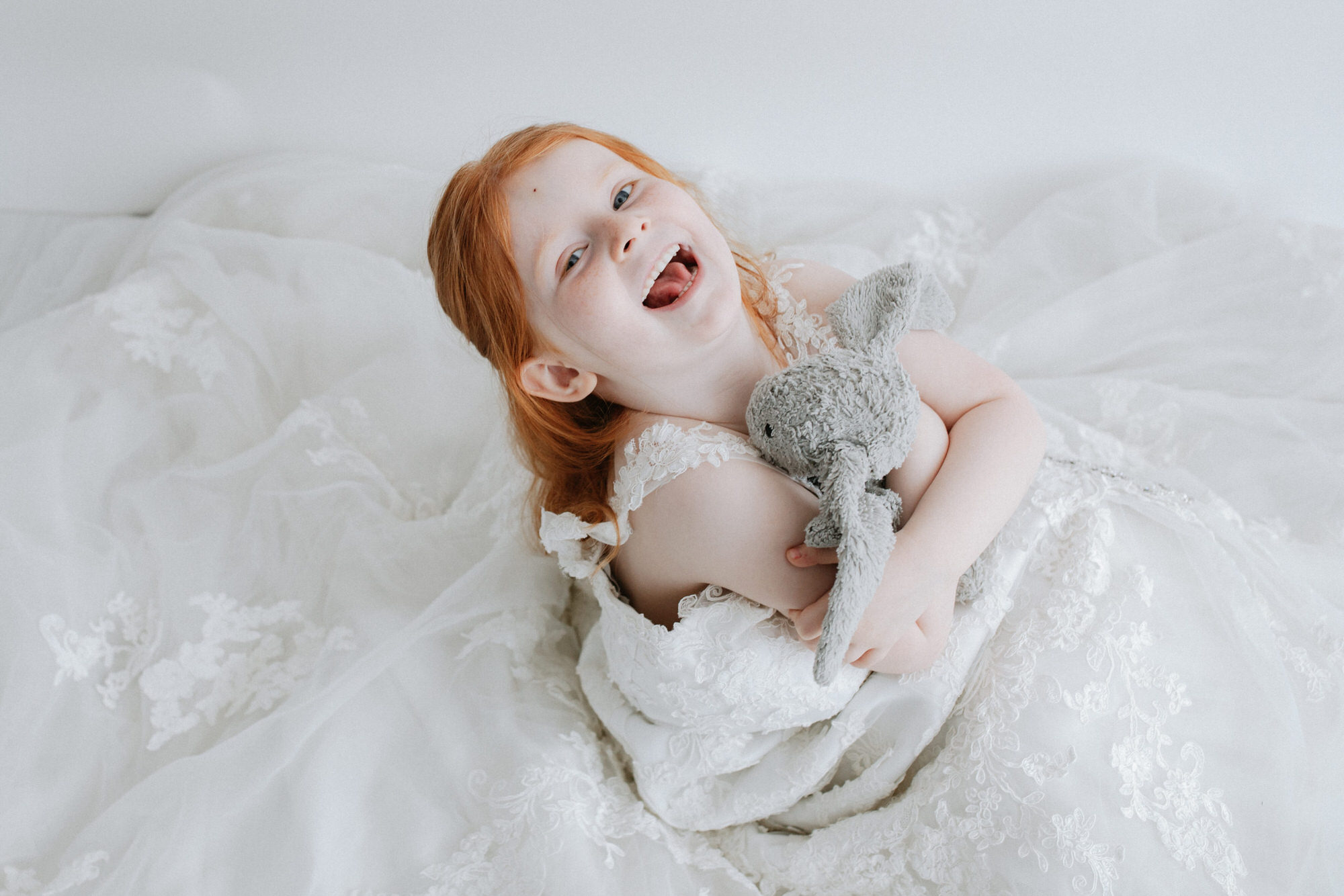 little girl looking up to the camera cuddling her teddy in her mummy's wedding dress at her shoot in Bexley
