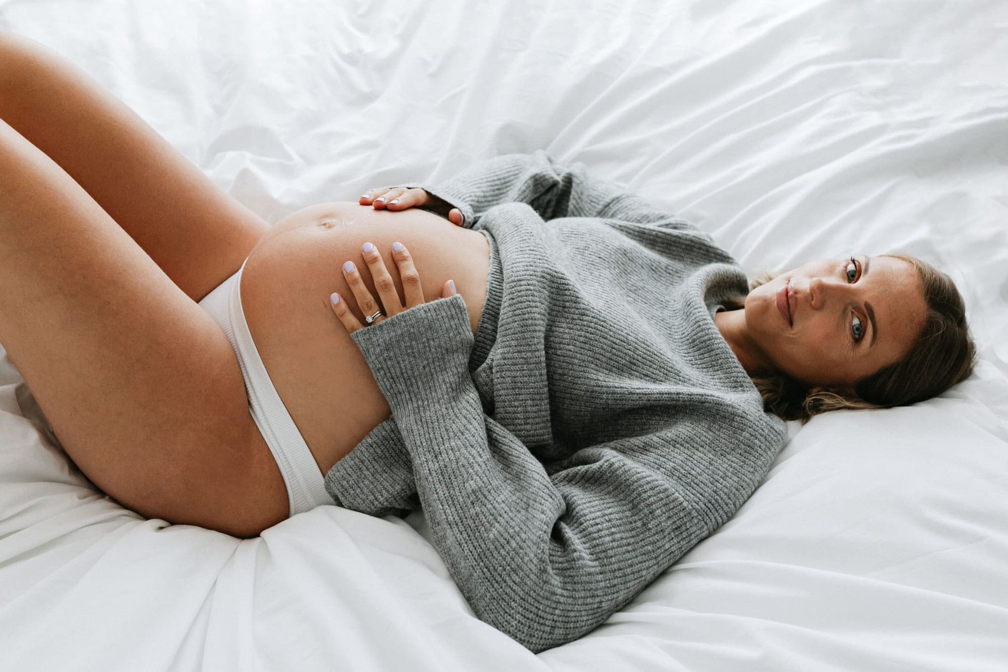 Mummy to be holding baby bump wearing oversized grey jumper laid on a bed at her kent maternity photoshoot in bexley