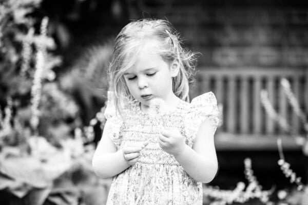 black and white image of a little girl deep in thought and looking at a dandelion clock at her kent family photoshoot in Sevenoaks