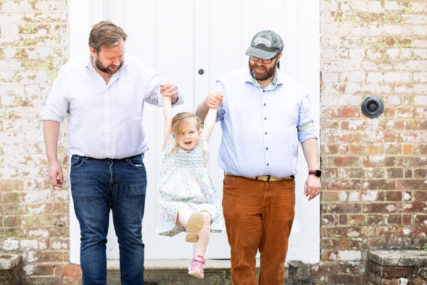 family of 3 with their little girl swinging between them infront of a rustic brick wall at their family photoshoot in Sevenoaks