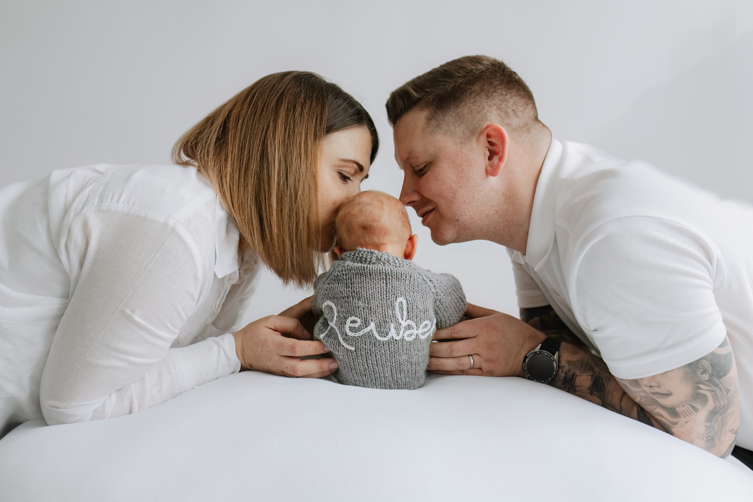 beautiful image of mummy and daddy leaning into their newborn baby sons kissing him on the head, he is wearing a hand knitted jumper with his name on at their kent newborn baby photoshoot in bexley