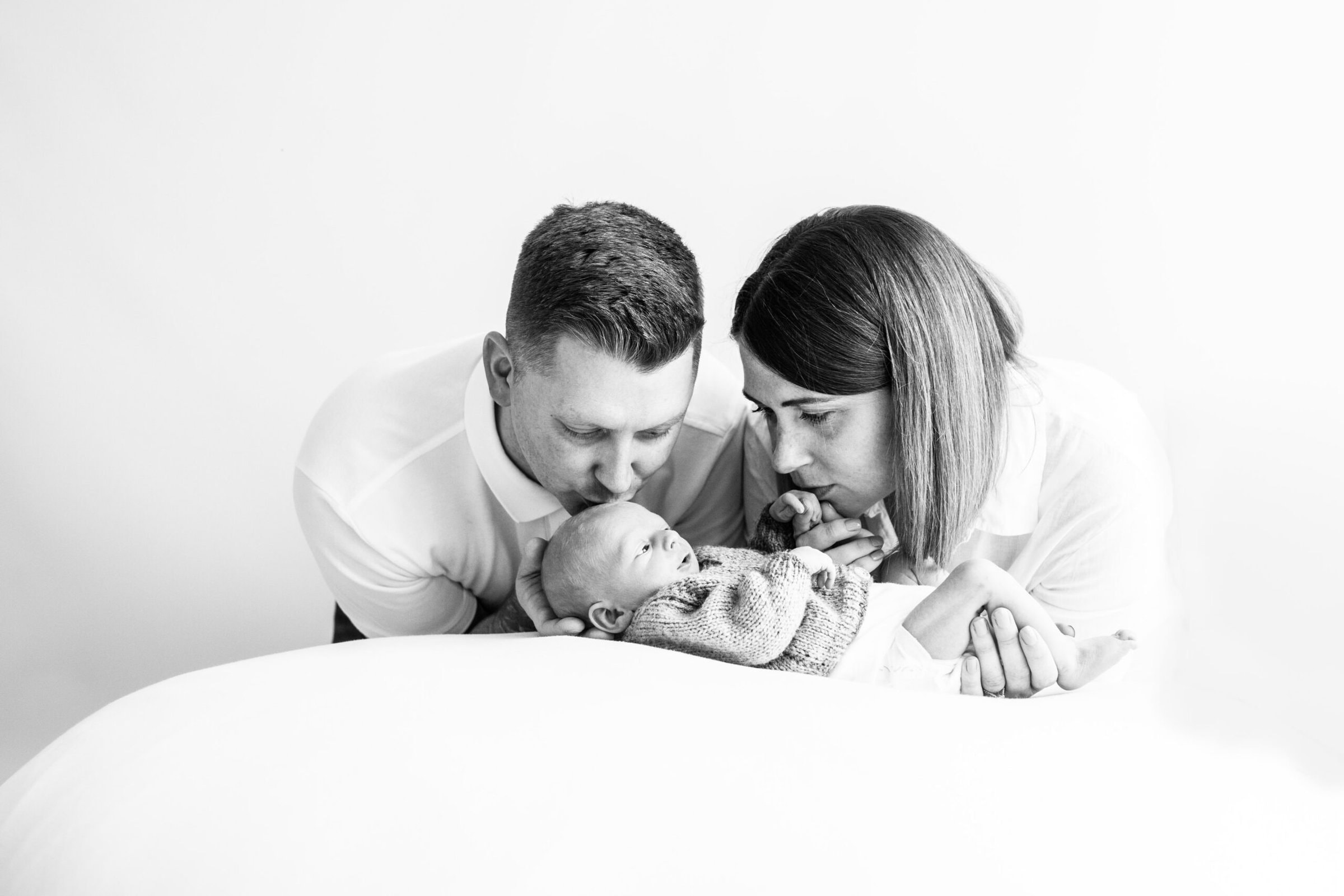 Mummy and daddy with their newborn son leaning in kissing him, photograph is black and white at their kent baby photoshoot in bexley