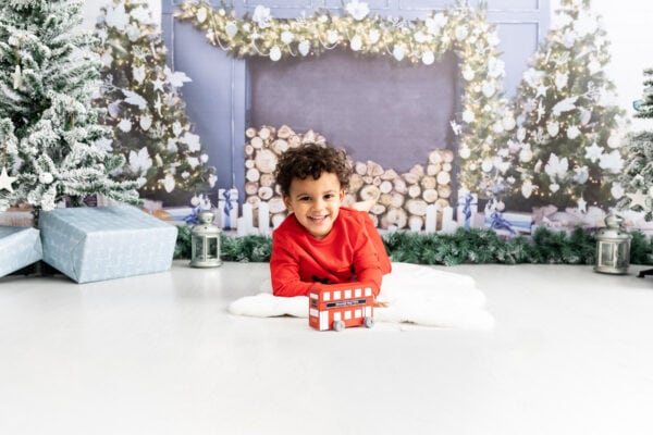 little boy playing with a red london bus in front of a blue christmas fire place at his christmas mini shoot