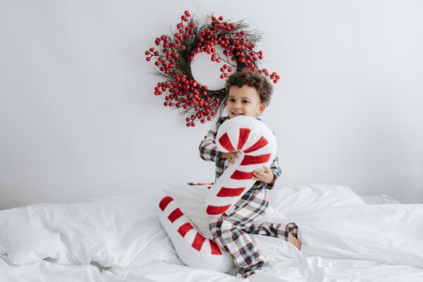 little boy jumping on a white bed in tartan christmas pyjamas cuddling a candy cane christmas cushion at his bexley christmas mini shoot