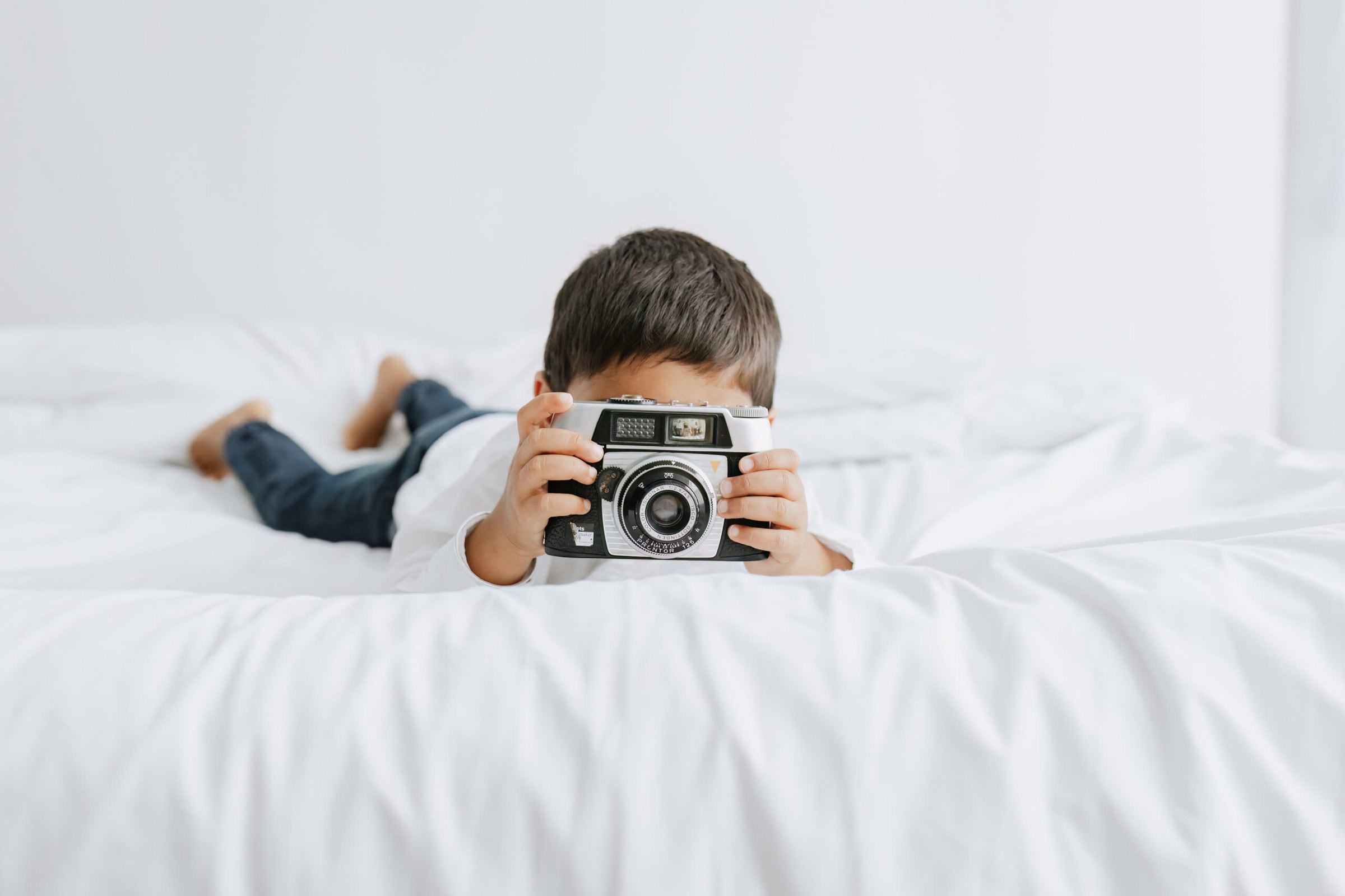 young boy with a vintage film camera sat on a white bed smiling at the camera at his bexley photoshoot in kent