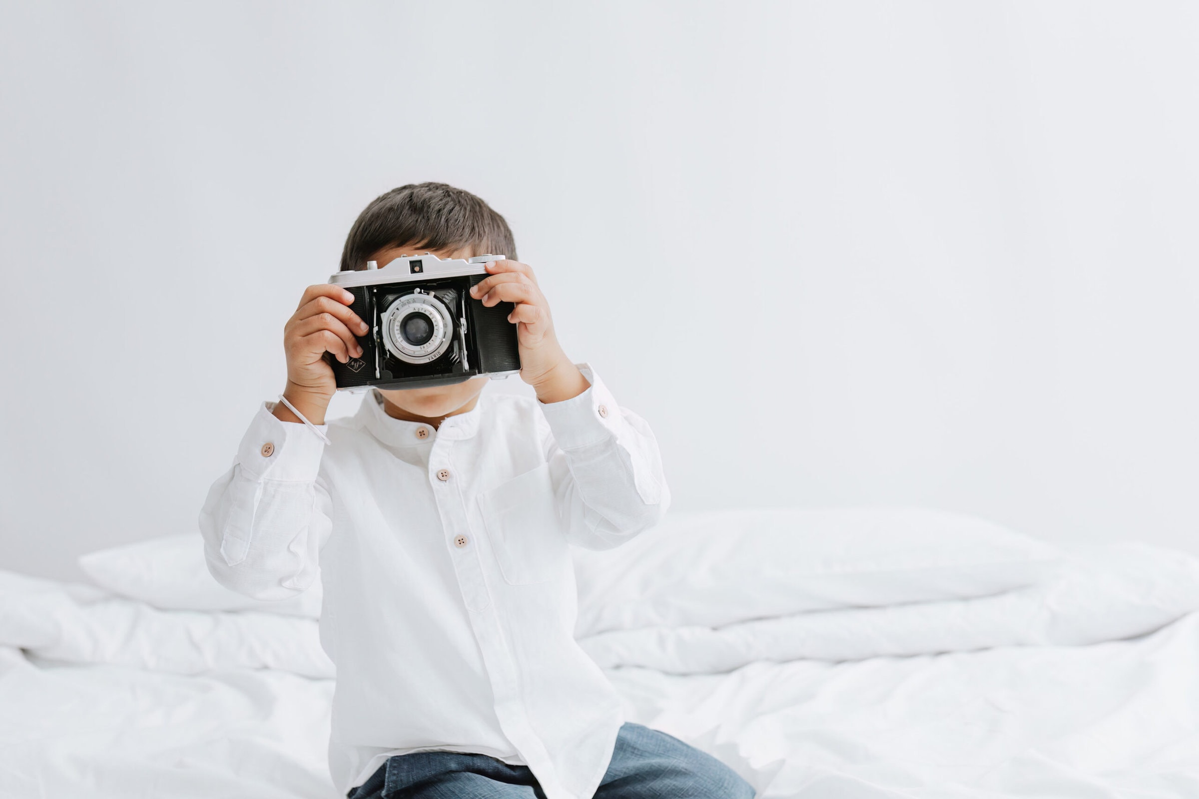 young boy with a vintage film Agfa camera sat on a white bed smiling at the camera at his bexley photoshoot in kent